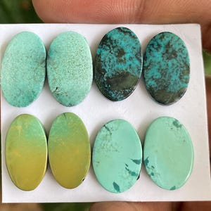May include: Six oval-shaped turquoise cabochons in various shades of green and blue. The cabochons are arranged in two rows of three on a white surface.