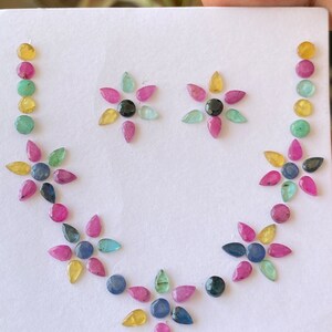 Puede incluir: Un conjunto de cuentas de piedras preciosas de colores dispuestas en un collar y dos pendientes. Las cuentas son en tonos de rosa, verde, azul y amarillo, con acentos negros. El collar está dispuesto sobre un fondo blanco.