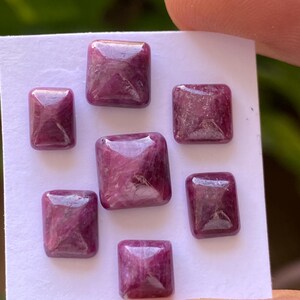 May include: Seven pink square-shaped gemstones with a faceted design. The gemstones are arranged in a random pattern on a white surface.