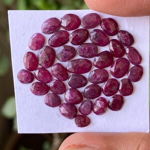 May include: A collection of numerous small, oval-shaped, faceted rubies with a deep red colour. The gemstones are arranged in a cluster on a square white paper backing, showcasing their translucent quality and reflective surfaces under light.