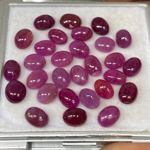 Puede incluir: Colección de gemas ovaladas pulidas en varios tonos de rojo y rosa. Las gemas se muestran en un pequeño recipiente gris abierto. Las piedras tienen una superficie lisa y brillante.