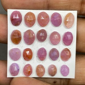 May include: Twenty oval gemstones in pink, peach, and purple hues. The faceted gems are arranged on a white card, showcasing their reflective surfaces. The collection highlights the gems' varying colours and clarity, suitable for jewellery making.