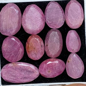 May include: A collection of faceted, oval-shaped pink gemstones displayed in a black tray. The gemstones vary slightly in color, ranging from light to deep pink. The facets catch the light, creating a sparkling effect. The gemstones are of various sizes.