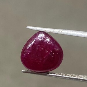 May include: A polished, heart-shaped ruby gemstone held by tweezers. The ruby is a deep, rich red colour with a smooth, reflective surface. The gemstone is a cabochon cut, ideal for jewellery.