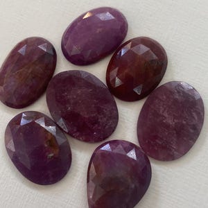 Puede incluir: Una colección de siete piedras preciosas facetadas de forma ovalada en color magenta intenso. Cada piedra presenta una superficie pulida con facetas geométricas que reflejan la luz, mostrando variaciones en la saturación del color y la textura.