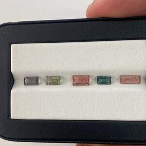 May include: Five rectangular gemstones in a black display case. The gemstones are various shades of green, pink, and gray.