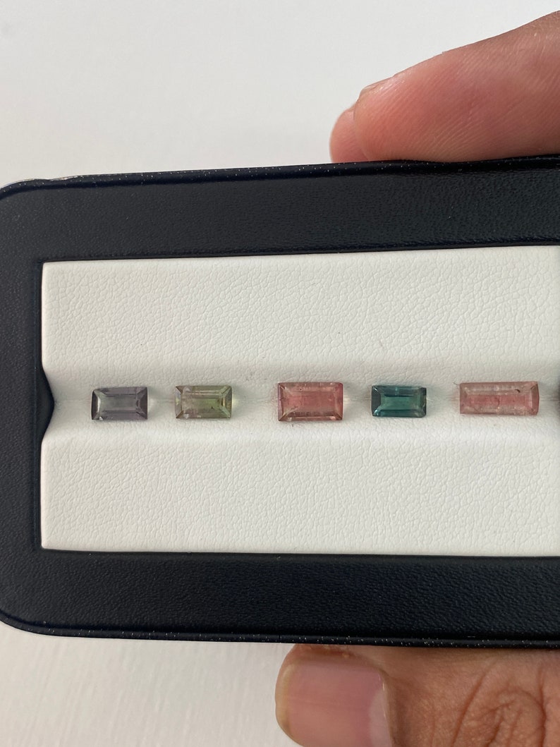 May include: Five rectangular gemstones in a black display case. The gemstones are various shades of green, pink, and gray.