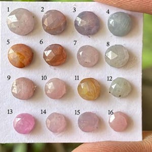 Puede incluir: Una colección de dieciséis piedras preciosas pequeñas, redondas y facetadas dispuestas en una cuadrícula de cuatro por cuatro sobre una tarjeta blanca. Cada piedra presenta un color único, incluyendo tonos de rosa, azul, naranja y morado, numeradas del uno al dieciséis.