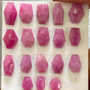 Puede incluir: Una colección de diecisiete piedras preciosas de rubí rosa facetadas dispuestas en cuatro filas sobre una tarjeta de presentación blanca. Cada gema presenta una forma geométrica distinta, incluyendo hexágonos y pentágonos.