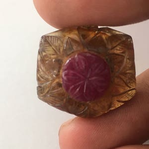 May include: A carved gemstone with a central, round, ruby-colored stone. The surrounding stone is a light amber color with carved leaf patterns. The gemstone is held between two fingers.