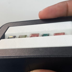 May include: A close-up of a black box with six small, rectangular gemstones. The gemstones are arranged in a row and are various colors, including green, pink, and black.