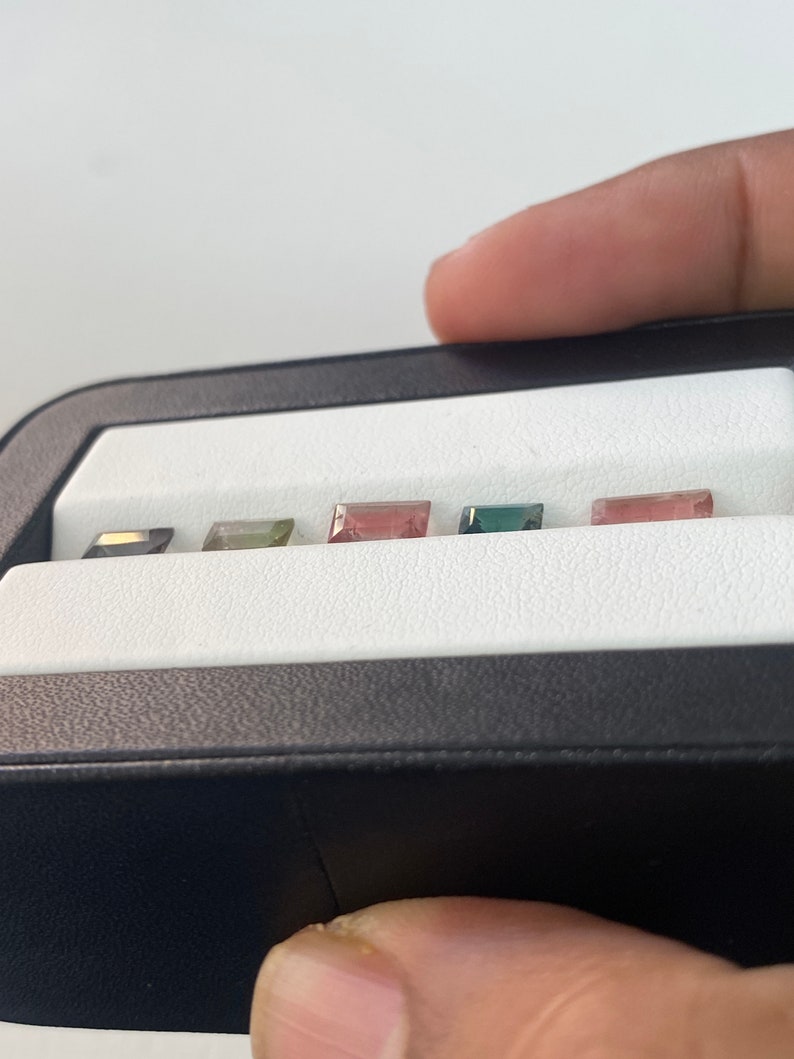 May include: A close-up of a black box with six small, rectangular gemstones. The gemstones are arranged in a row and are various colors, including green, pink, and black.