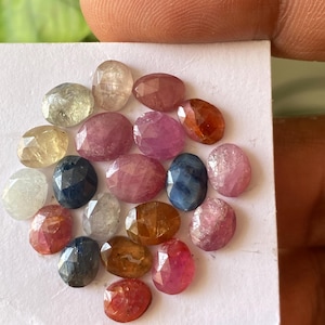 Peut inclure: Une collection de pierres précieuses de forme ovale, de différentes couleurs, notamment rose, bleu, orange et jaune. Les pierres sont présentées sur une surface blanche, mettant en valeur leurs facettes et leur clarté. Les gemmes sont tenues par un doigt.