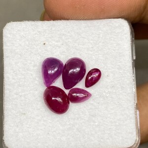 May include: Five loose gemstones in a white box. The gemstones are various shades of pink and purple. The gemstones are in a variety of shapes, including oval and pear.