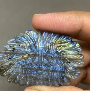 May include: A carved labradorite gemstone in a floral design. The stone features a detailed carving of flowers and leaves, with iridescent blue, green, and gold flashes. The gemstone is held in a hand.