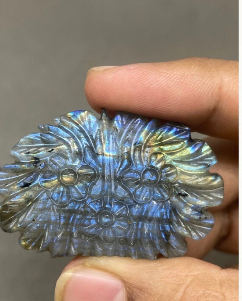 May include: A carved labradorite gemstone in a floral design. The stone features a detailed carving of flowers and leaves, with iridescent blue, green, and gold flashes. The gemstone is held in a hand.