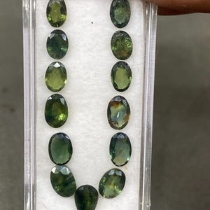 Peut inclure: Un étui en plastique transparent contient une collection de pierres précieuses ovales et en forme de poire. Les pierres présentent des nuances de vert et d'olive, avec des variations de clarté et de saturation des couleurs. L'arrangement met en valeur les facettes et les teintes naturelles.