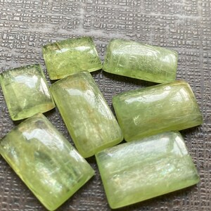 May include: Seven light green, rectangular gemstones with rounded corners are arranged on a textured surface. The stones have a translucent quality, with visible internal structures and a subtle shimmer. The overall color is a soft, pale green.