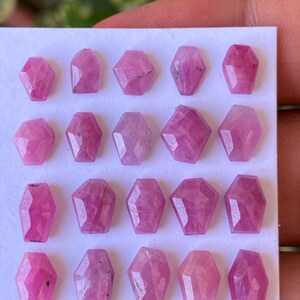 May include: A collection of twenty pink gemstones, each cut into a unique coffin shape. The gems are arranged in rows on a white surface, showcasing their faceted surfaces and varying shades of pink.