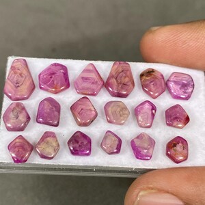 May include: A collection of pink and purple gemstones with various geometric shapes. The stones are displayed on a white surface, showcasing their natural patterns and colors. The gemstones vary in size and have a polished appearance.