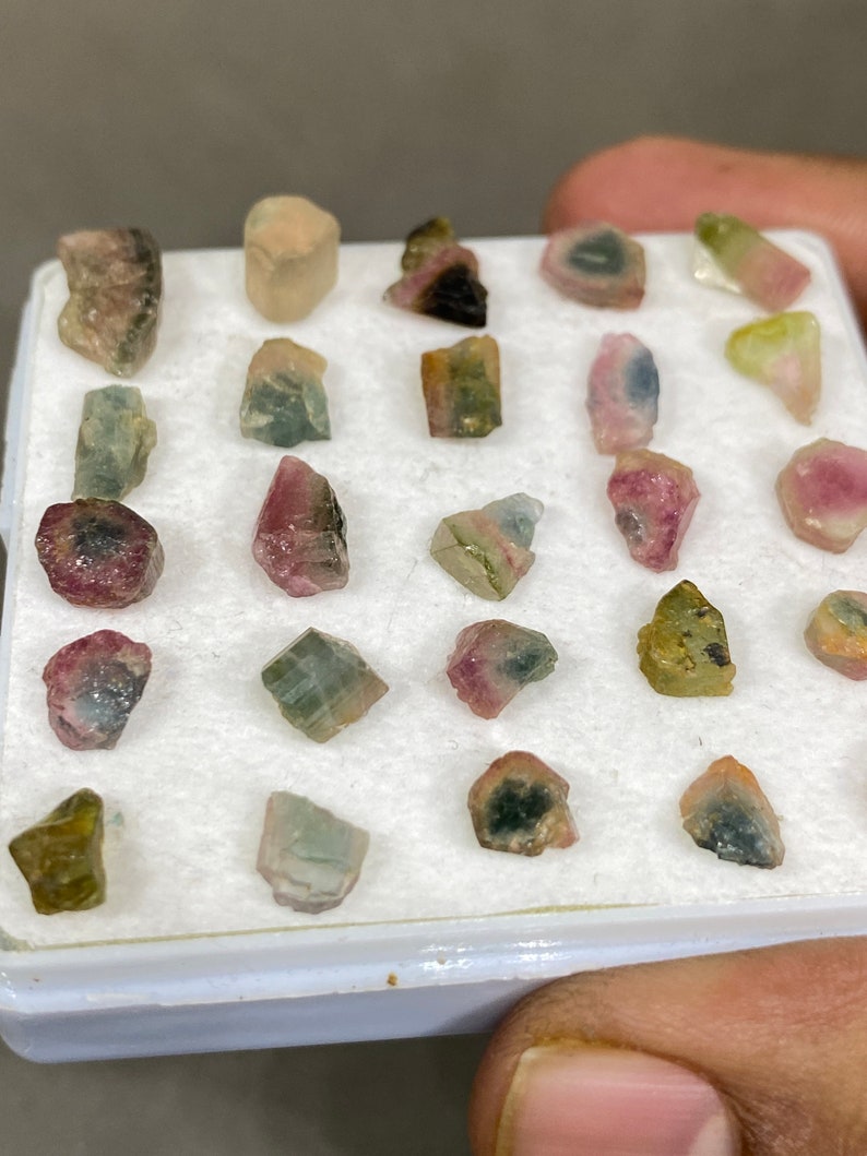 May include: A collection of 20 small, colourful, raw tourmaline crystals in a white box. The crystals are various shades of pink, green, and black.