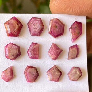 Op de afbeelding: Een verzameling gefacetteerde, roze edelstenen met verschillende geometrische vormen. De edelstenen zijn gerangschikt op een wit oppervlak, waarbij hun gepolijste oppervlakken en interne patronen worden benadrukt. De kleur varieert van licht- tot dieproze, sommige vertonen interne adering.