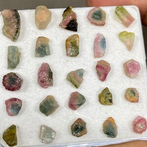 May include: A collection of 20 small, colourful, raw, rough, and uncut gemstones. The gemstones are arranged in a white foam tray. The gemstones are various shades of pink, green, blue, and black.