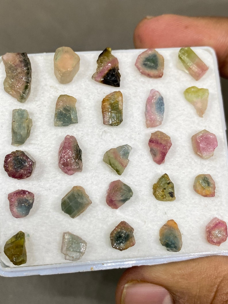 May include: A collection of 20 small, colourful, raw, rough, and uncut gemstones. The gemstones are arranged in a white foam tray. The gemstones are various shades of pink, green, blue, and black.