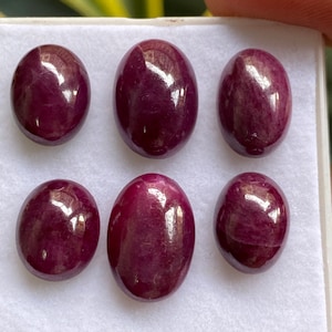 May include: Six oval-shaped, deep red gemstones with a polished finish. The gemstones are displayed in a white box.