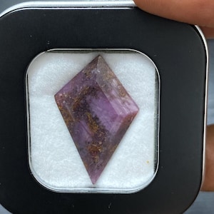 May include: A faceted, diamond-shaped gemstone with a deep purple hue and brown inclusions. The gem is set in a black square case with a white interior, held by a person's hand. The stone's facets catch the light, showcasing its unique color and pattern.