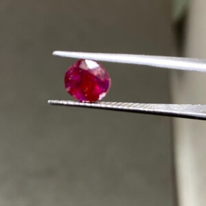 May include: A faceted, oval-shaped ruby gemstone held by tweezers. The ruby is a deep, saturated red color, with visible internal inclusions. The tweezers are silver-colored, and the background is out of focus.