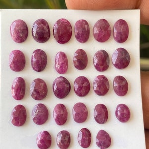 May include: A collection of oval-shaped, faceted ruby gemstones. The rubies are a deep, rich red color, with variations in tone and clarity. They are arranged on a white surface, showcasing their individual shapes and cuts. The gemstones are ready for jewelry making.