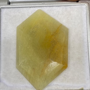 Peut inclure: Une pierre précieuse hexagonale facettée, de couleur jaune pâle avec de subtiles inclusions orange et noires. La gemme est placée dans un plateau argenté, mettant en valeur ses surfaces polies et ses variations de couleur uniques. La pierre est une matière première naturelle.