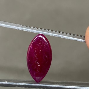 Op de afbeelding: Een dieprode edelsteen in marquise-vorm wordt vastgehouden door een pincet. De gepolijste steen heeft een glad oppervlak en een puntige, langwerpige vorm. De kleur van de edelsteen is een rijk, verzadigd rood, met subtiele kleurvariaties.