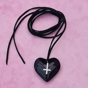 Puede incluir: Un colgante negro en forma de corazón con un dije de cruz plateada cuelga de un cordón negro. El colgante es brillante y liso, y el cordón está en bucle. El fondo es de color rosa liso.
