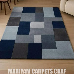 Peut inclure: Un tapis rectangulaire présentant un motif géométrique de blocs imbriqués dans des tons bleu marine, gris anthracite et bleu clair. Le tapis affiche un design à grille moderne avec des rectangles de tailles variées et le texte MARIYAM CARPETS CRAF.