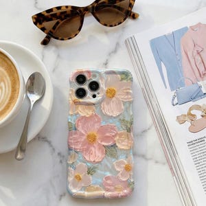 Puede incluir: Funda de teléfono con estampado floral y diseño texturizado en rosa pastel, amarillo y azul. Gafas de sol carey y una taza de café con arte latte también son visibles.
