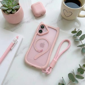 Peut inclure: Un étui de téléphone rose avec un design d'oreille de chat et une dragonne assortie. L'étui est sur une surface blanche avec une plante succulente dans un pot rose, un stylo rose, des écouteurs sans fil roses et une tasse de café.