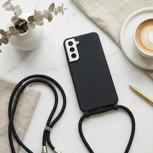 May include: A black phone case with a matching black lanyard. The phone case has a camera cutout and is lying on a marble surface. A cup of coffee and a pen are also on the surface.