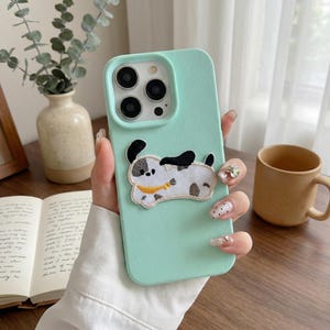 May include: A mint green phone case featuring a playful embroidered dog design. The case has a smooth texture and a camera cutout. The dog has black ears and spots, with a yellow collar.