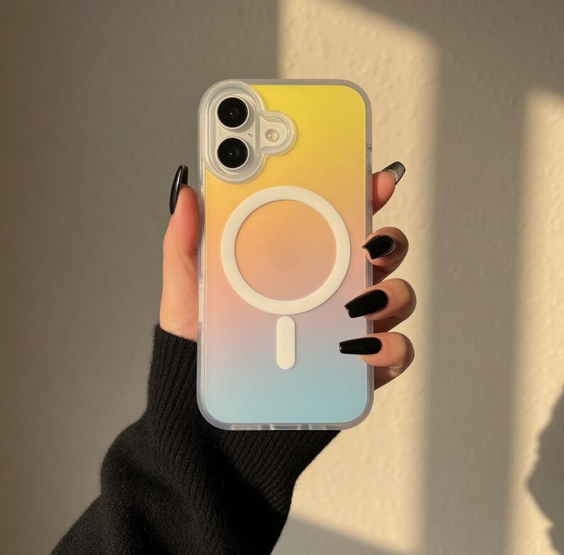 May include: A clear phone case with a gradient design, transitioning from yellow to orange to blue. The case features a white circular MagSafe charger and a camera cutout. The phone case is held by a person.