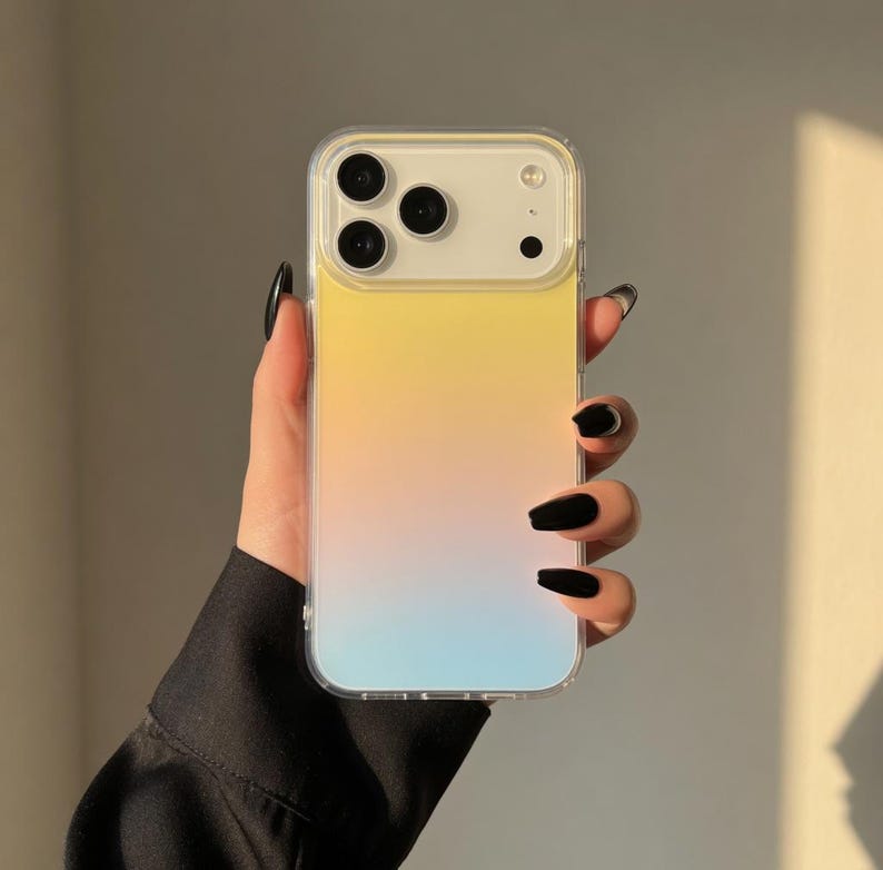May include: A clear phone case with a gradient design transitioning from yellow to pink to blue. The case has a clear border and a camera cutout. The phone case is held in a hand with black nail polish.