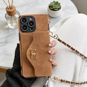 Op de afbeelding: Een bruine telefoonhoes met een portefeuille-achtige flap en een gouden sluiting. De hoes heeft een kettingriem met gouden en bruine schakels. De telefoonhoes is ontworpen om een telefoon en kaarten in op te bergen.