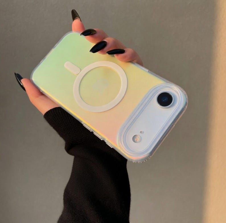 May include: Iridescent phone case with a white circular MagSafe charger. The case has a clear border and a camera cutout. The case reflects yellow, green, and pink hues. The phone case is designed to protect the phone.