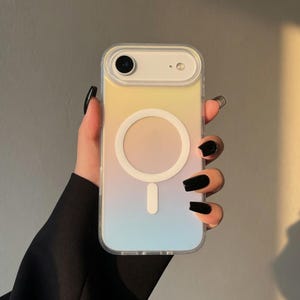 May include: A clear phone case with a gradient design of yellow, peach, and blue. The case has a white circular magnetic charging area and a white rectangular charging port. The phone case is held in a hand with black nail polish.