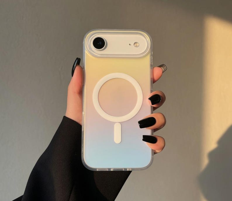 May include: A clear phone case with a gradient design of yellow, peach, and blue. The case has a white circular magnetic charging area and a white rectangular charging port. The phone case is held in a hand with black nail polish.