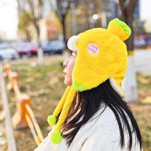 May include: A bright yellow winter hat with ear flaps and a rounded top, accented with lime green trim. The hat features a small, round patch that reads "100% LUCK". The hat's ear flaps have yellow tassels with lime green pom-poms.