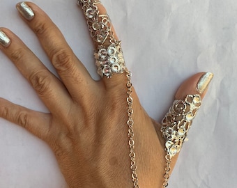 May include: A silver ring with a pointed design and a chain connecting to a second ring. The ring is adorned with clear rhinestones.