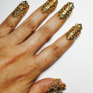 May include: Gold-colored filigree nail covers adorn a hand, showcasing intricate openwork designs. The nail covers are shaped to fit each nail, adding a touch of elegance and unique style. The ornate nail art is a statement piece.