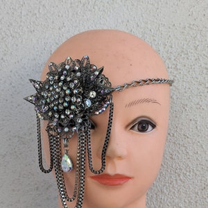 May include: A decorative eye patch featuring a dark gray floral design with iridescent rhinestones and dangling chains. The eye patch is attached to a silver chain that goes around the head. A teardrop-shaped iridescent crystal hangs from the chains.